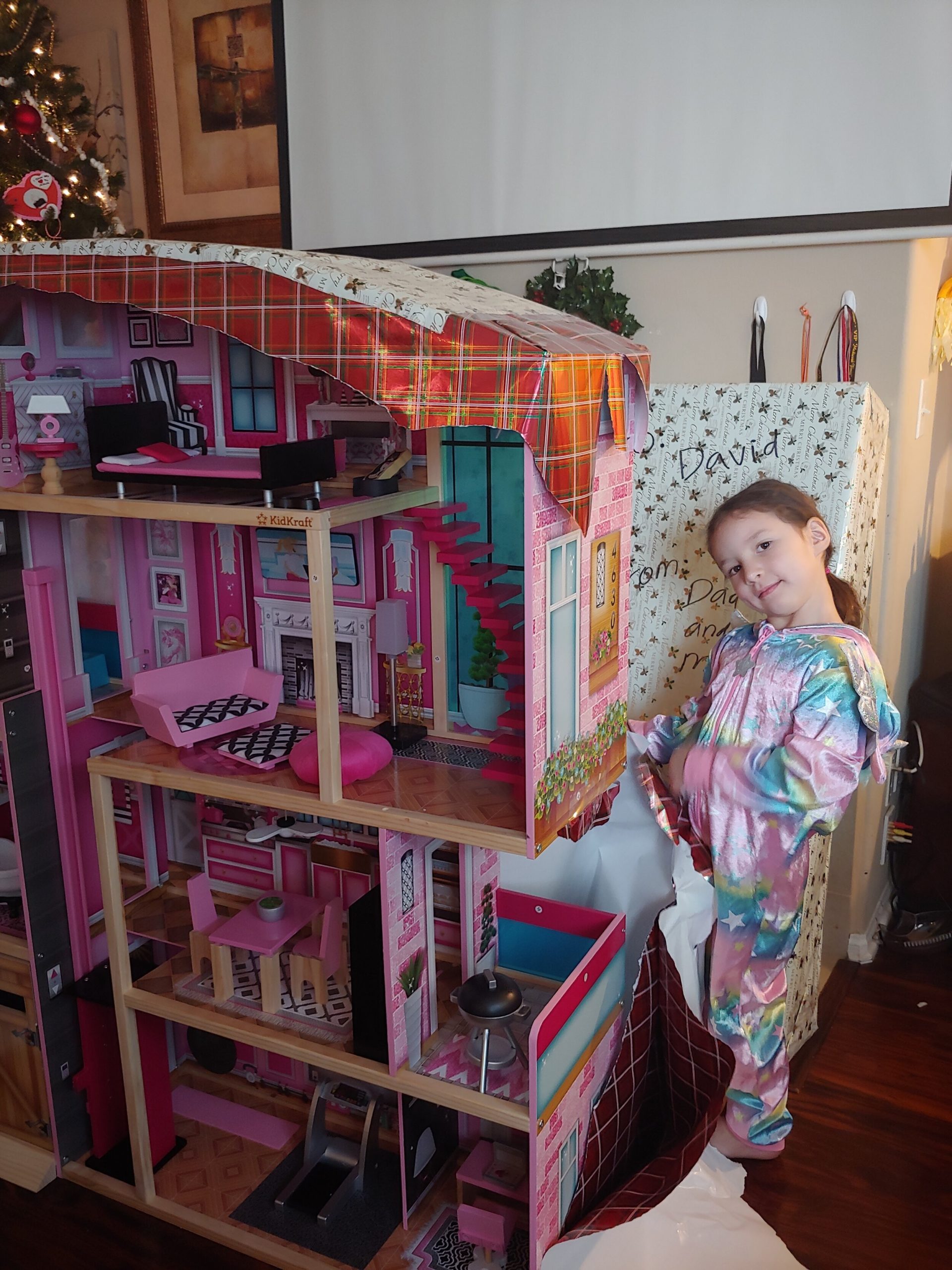 a little girl unwrapping a Christmas present that is a doll house as tall as her